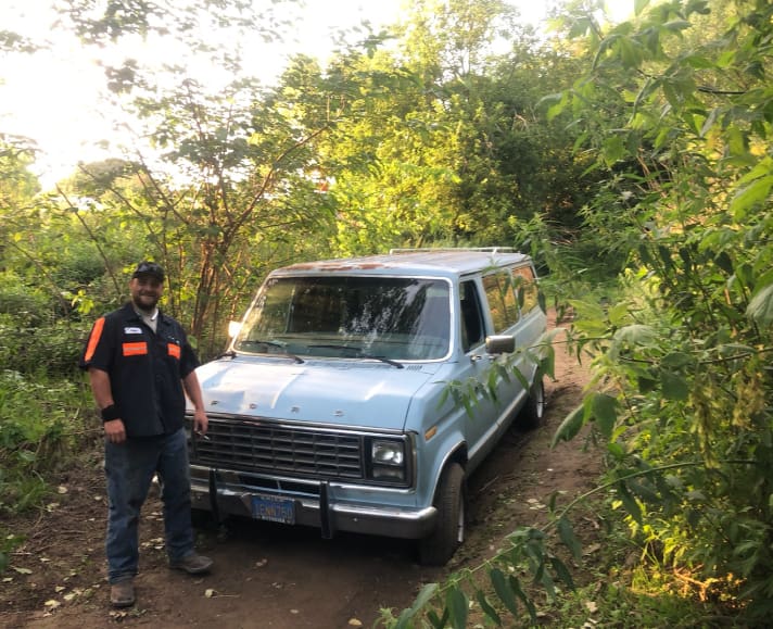 A man standing next to an old van in the woods.