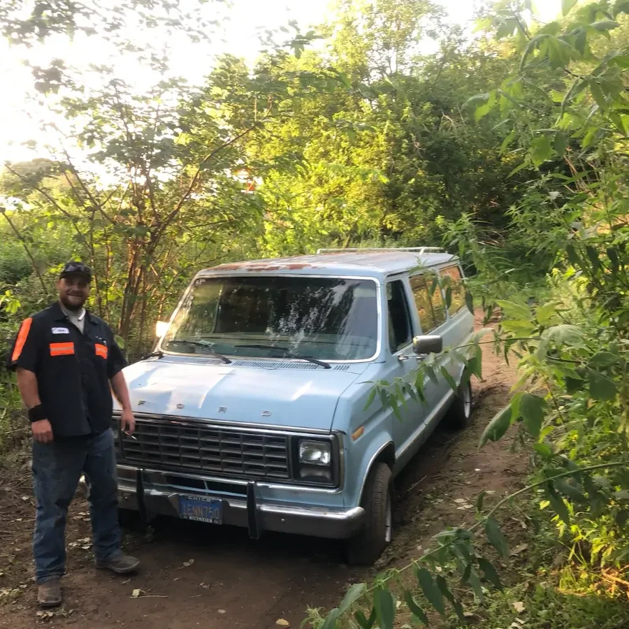 A man standing next to an old van in the woods.